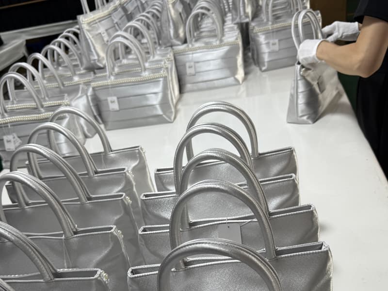 Rows of shiny silver satin evening tote bags being checked by QC staff with white gloves